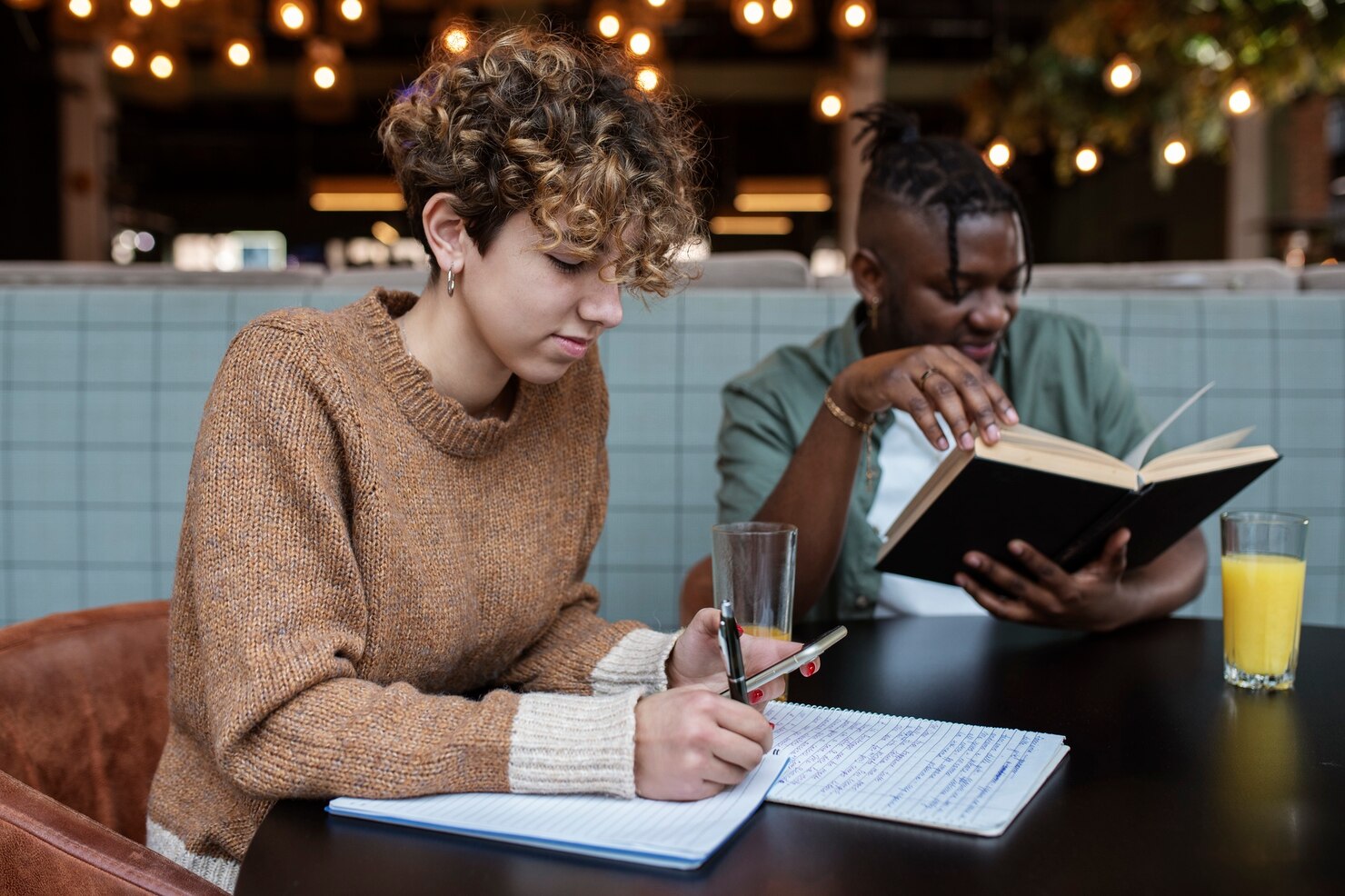 Two people sitting at a table in a warmly lit cafe or restaurant, one writing notes in a notebook with a pen and the other reading a book. Both have drinks on the table, one with water and the other with orange juice. The atmosphere appears focused and collaborative.