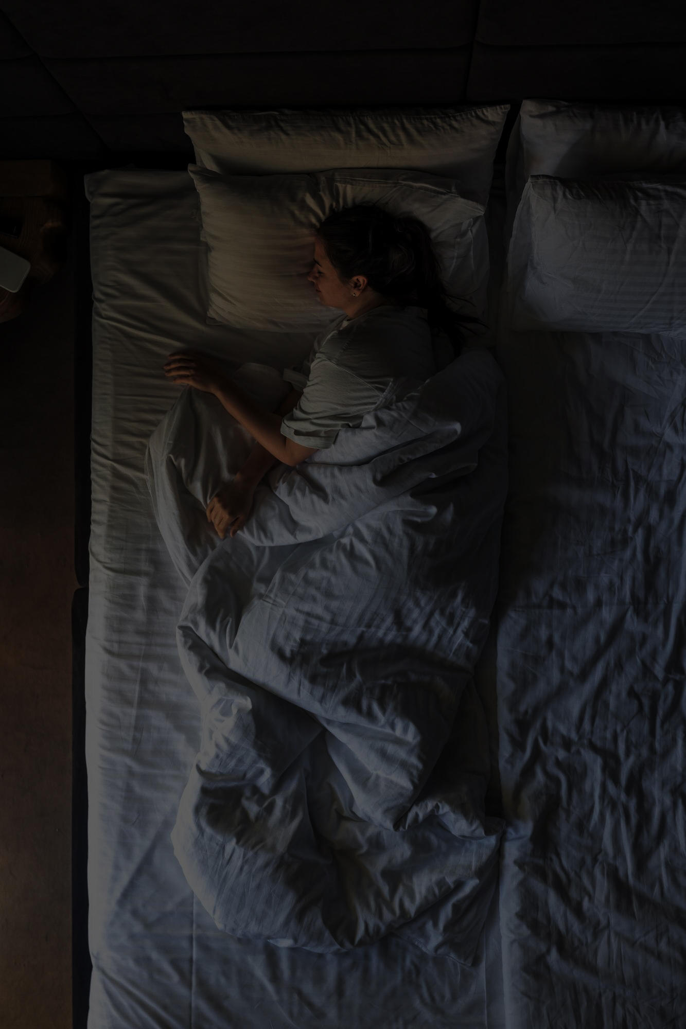 Person lying on a bed under a white comforter, resting on their side with one arm bent and the other extended. The room is dimly lit, creating a calm and peaceful nighttime atmosphere. The bedding is neatly arranged, and the overall environment feels quiet and restful.