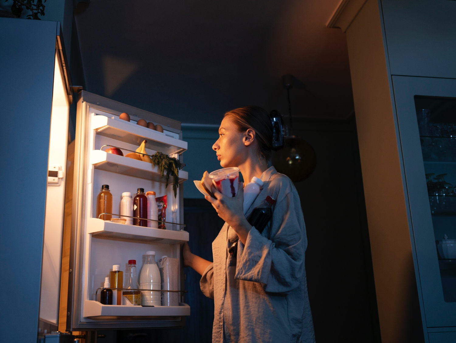 A person stands in front of an open refrigerator at night, holding a container of food and looking inside. The kitchen is dimly lit with the refrigerator light illuminating shelves stocked with eggs, bottles, and various groceries. The atmosphere feels quiet and contemplative, suggesting a late-night snack or search for something to eat.