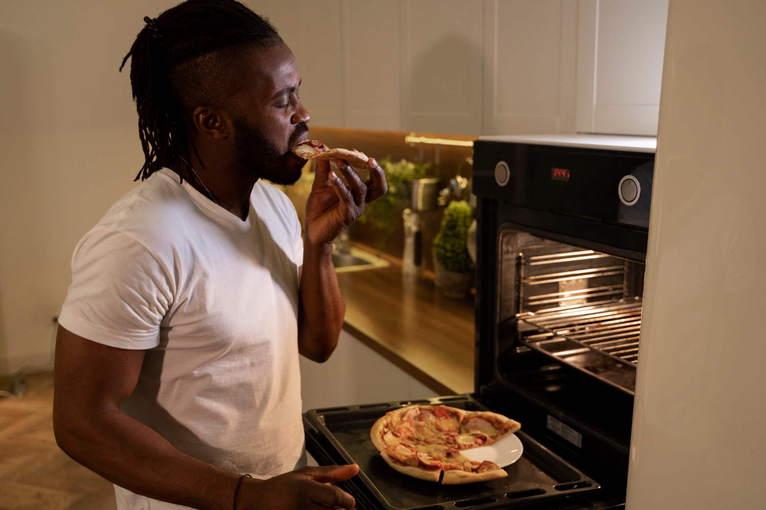 A man eats pizza straight from a tray in the kitchen late at night, illustrating how a heavier meal close to bedtime can become the other extreme that disrupts sleep.