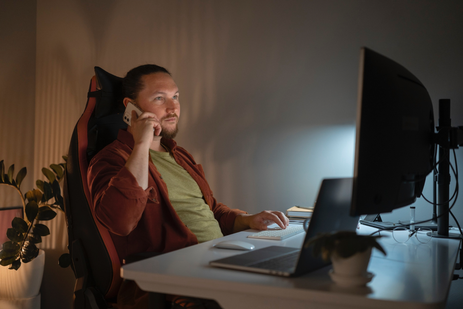 Person sitting at a desk in a modern home office, talking on a smartphone, with a computer monitor, laptop, keyboard, mouse, and a potted plant visible on the desk. The room is softly lit with a warm ambiance.