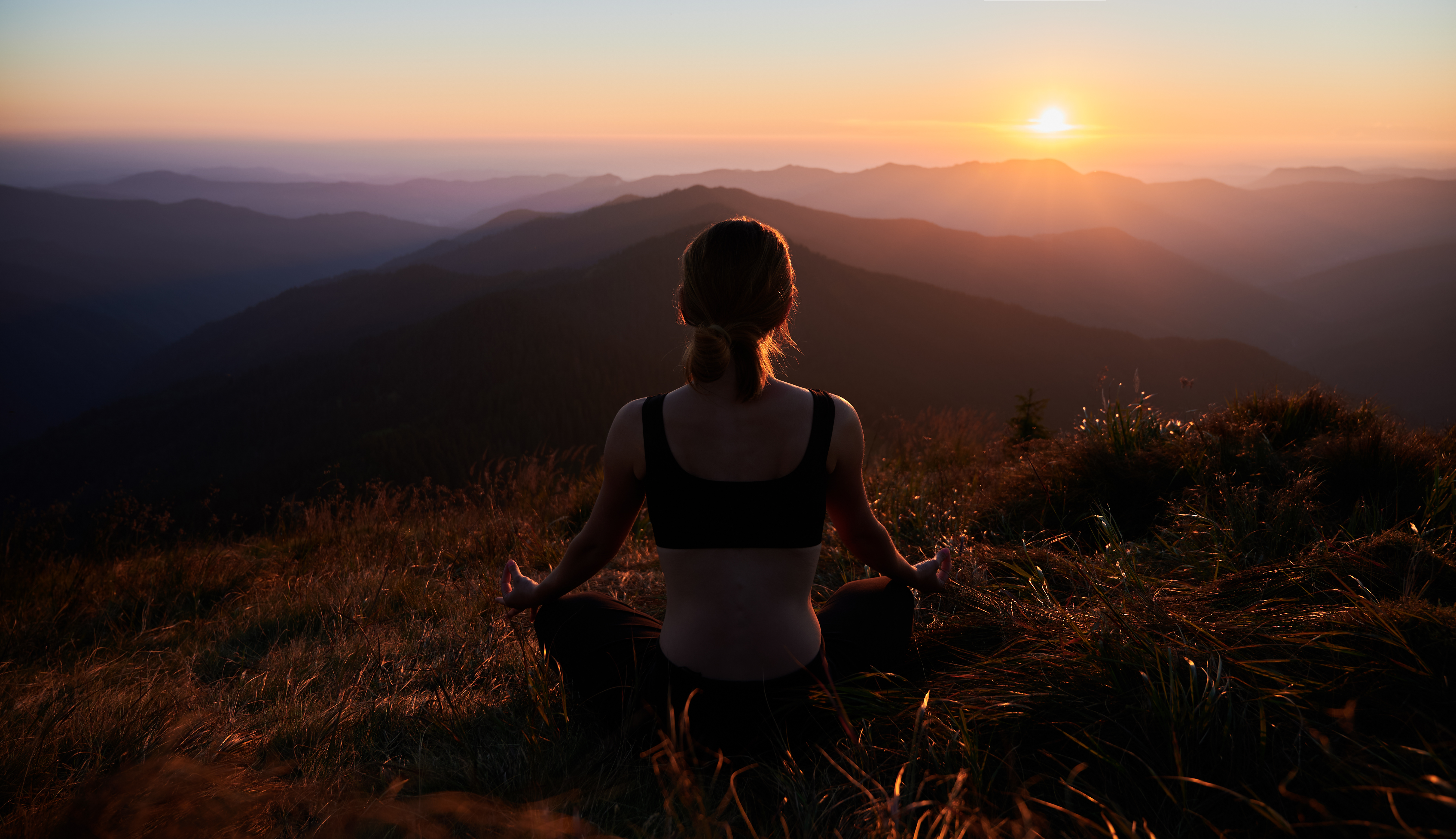 A woman sits cross-legged on a grassy mountain slope at sunrise, facing away and meditating with hands resting on her knees. The landscape features rolling hills and distant mountain ranges bathed in warm golden light. The scene feels peaceful, serene, and contemplative, evoking a sense of calm and connection with nature.