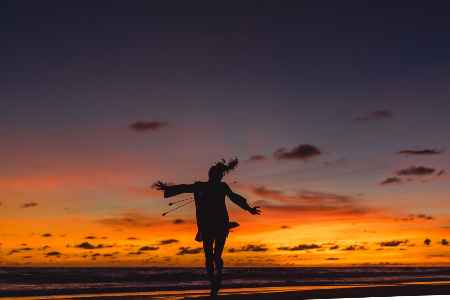 Silhouette of a person dancing or jumping with arms outstretched on a beach at sunset. The sky is filled with vibrant orange, yellow, and purple hues, with scattered clouds and the ocean visible in the background. The scene feels joyful, free, and energetic, evoking a sense of happiness and connection with nature.