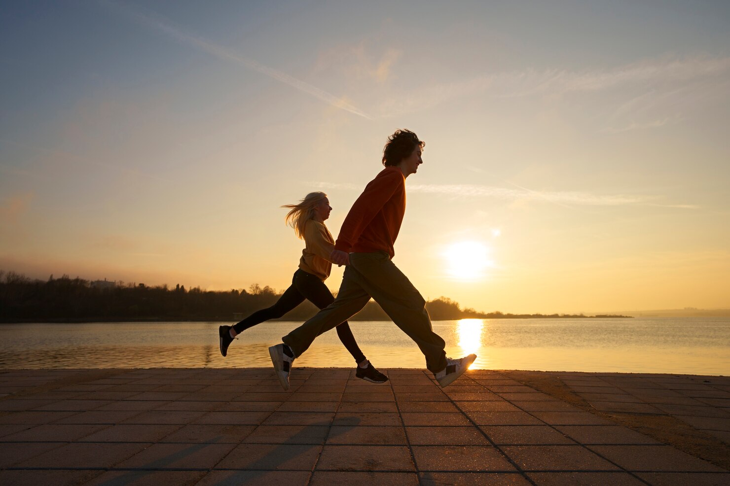 Two people run hand in hand along a paved walkway beside a body of water at sunset. The sun is low on the horizon, casting long shadows and bathing the scene in warm golden light. The background features calm water, distant trees, and a clear sky with wispy clouds. The mood feels energetic, joyful, and free, suggesting a sense of togetherness and enjoyment of the outdoors.