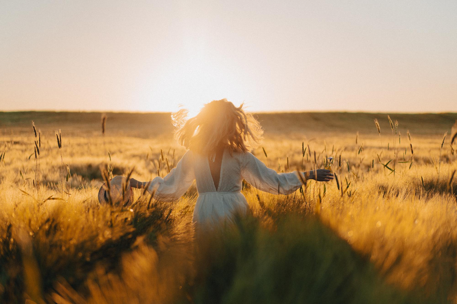 A woman in a white dress walks through a golden wheat field at sunset, her back to the camera and arms outstretched, gently brushing the tops of the tall grass. The sun is low on the horizon, casting a warm, glowing light across the landscape. The scene feels peaceful, free, and joyful, evoking a sense of connection with nature and serenity.