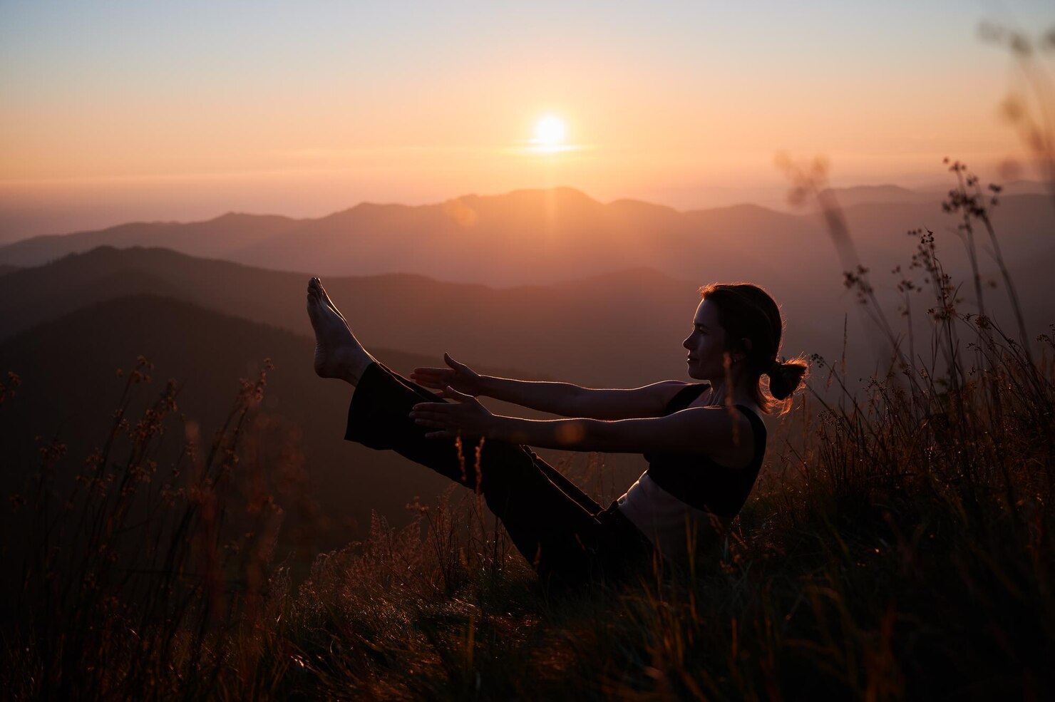 Person practicing yoga outdoors at sunset, seated on a grassy hillside with legs and arms extended in a V shape. The background features rolling mountain ranges and a glowing sun low in the sky, casting a warm, tranquil light over the scene. The overall mood is peaceful and reflective, emphasizing a connection with nature.