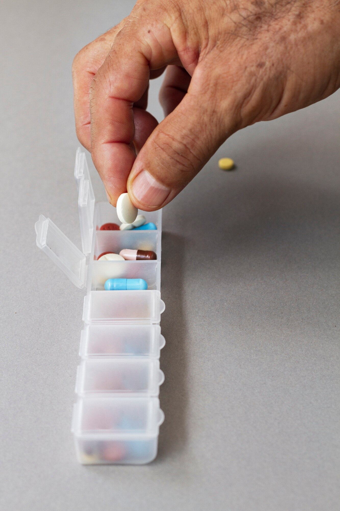 Hand placing a white oval pill into a transparent weekly pill organizer filled with various colored capsules and tablets. The background is a plain light gray surface, and the scene conveys a sense of routine, care, and attention to health management.