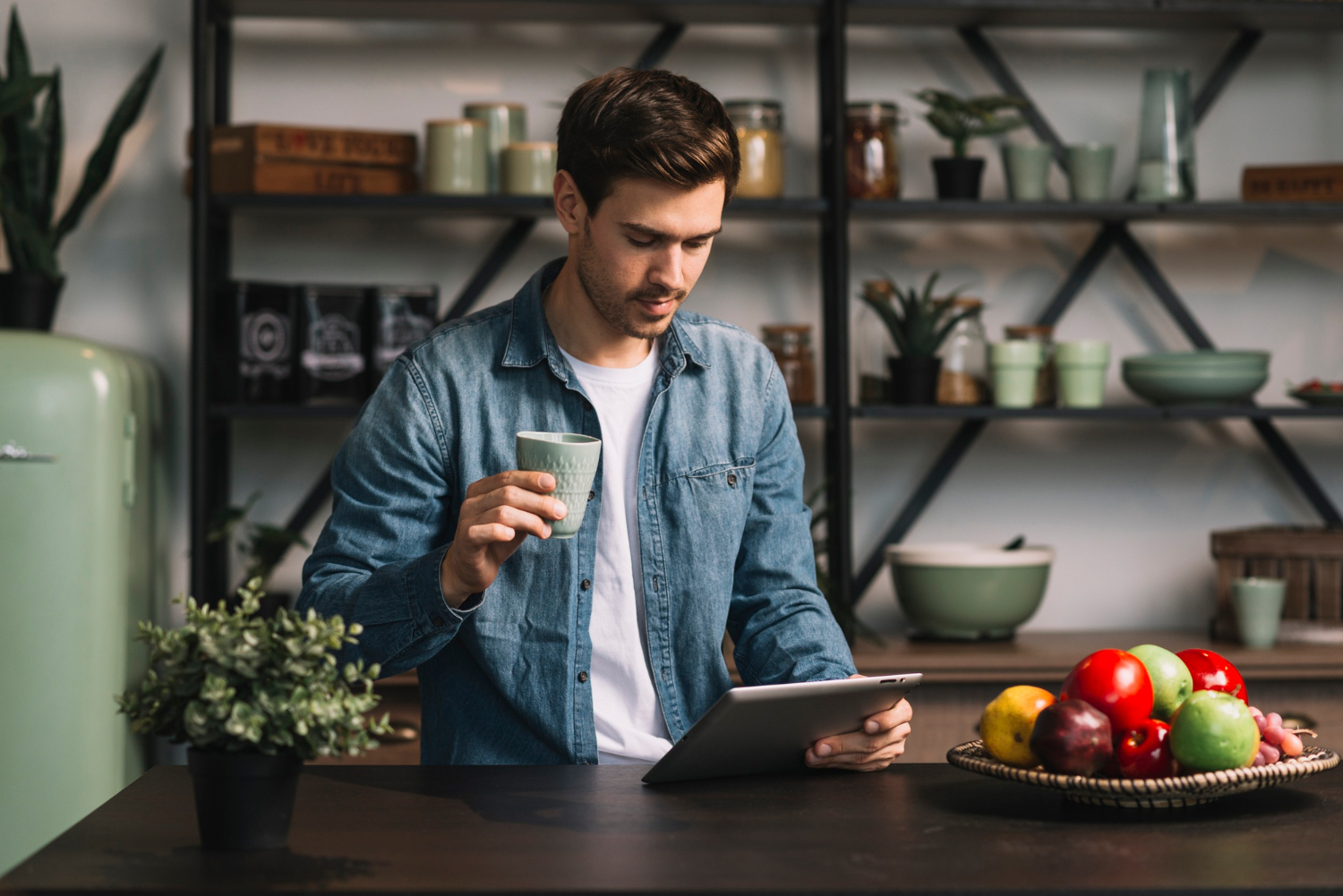 A man stands in a kitchen holding a mug and looking at a tablet, with a bowl of fruit on the counter and shelves of dishes behind him.
