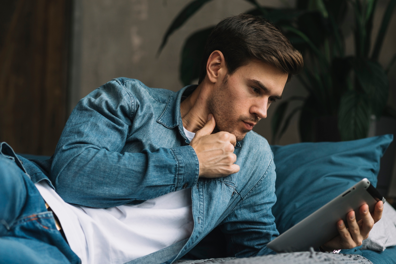 A man lies on a bed in casual clothes, holding a tablet and looking at the screen while one hand rests near his throat. Blue bedding and a softly lit bedroom setting frame the scene.