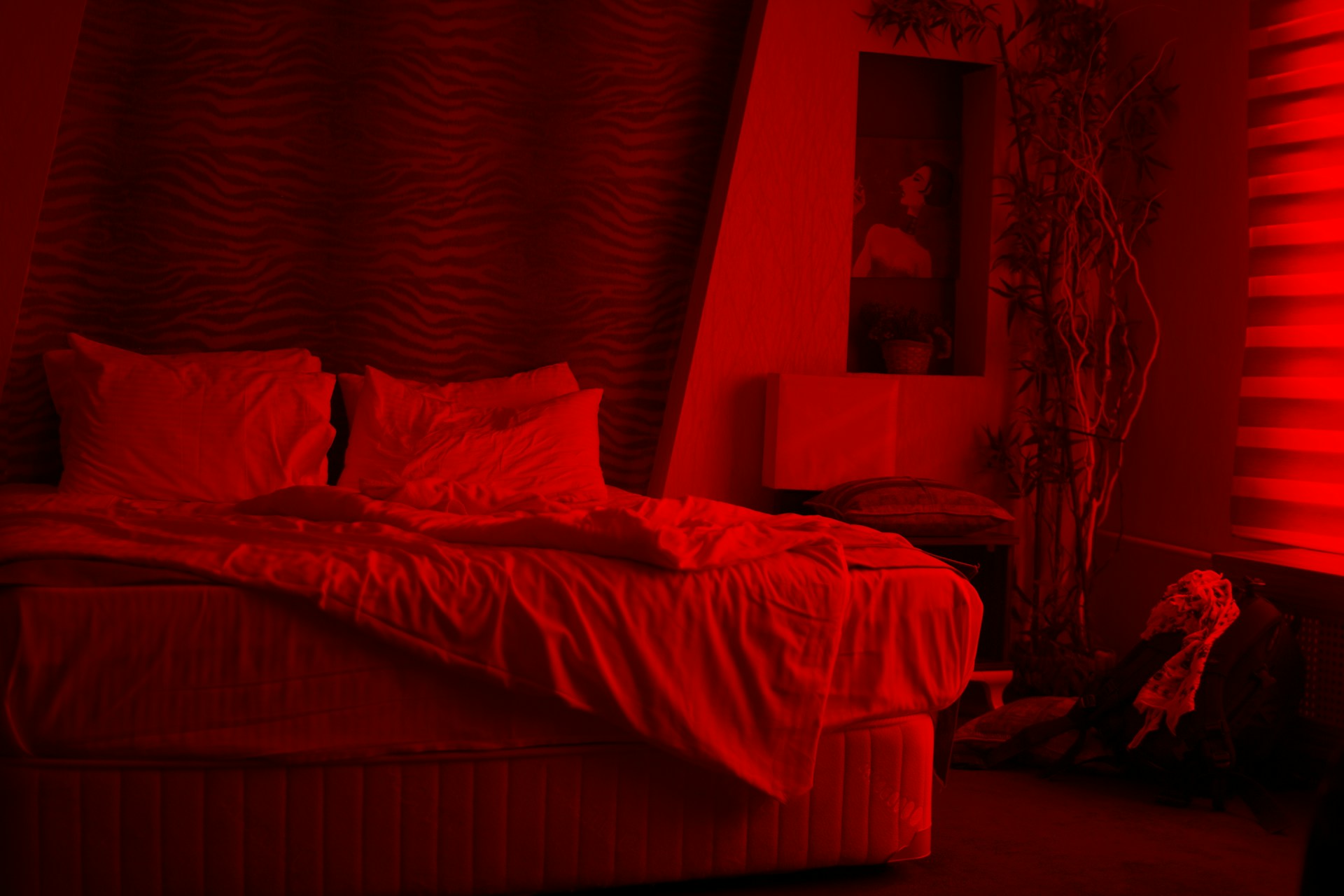 A person sits on a bed in a dark room, facing away and wrapped in a blanket, with only their silhouette visible in soft light. The bed has rumpled sheets and pillows, and the overall atmosphere feels quiet, introspective, and somber, suggesting solitude or contemplation at night.