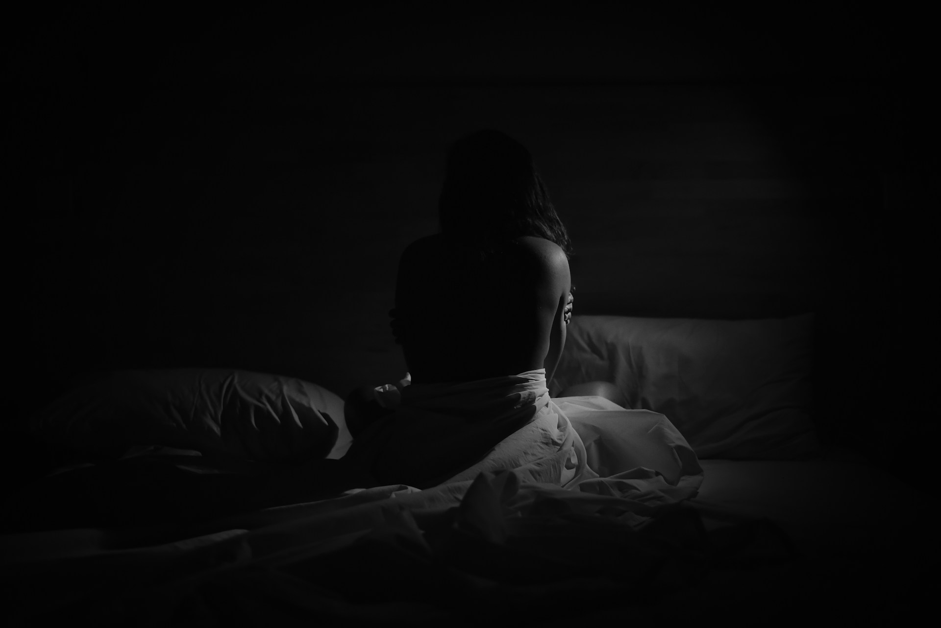 A dimly lit bedroom bathed in red light features an unmade bed with rumpled sheets and pillows. The room includes a patterned wall, a small table with a cushion, a potted plant, and a backpack with clothing draped over it. The atmosphere feels moody and mysterious, suggesting a late night or early morning setting.