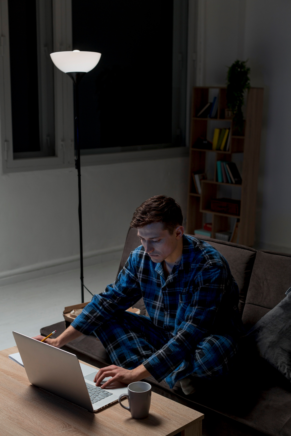 A man in blue plaid pajamas sits on a couch at night working at a laptop beside a mug and a bright floor lamp, showing how light and task-focused activity can keep the brain engaged.