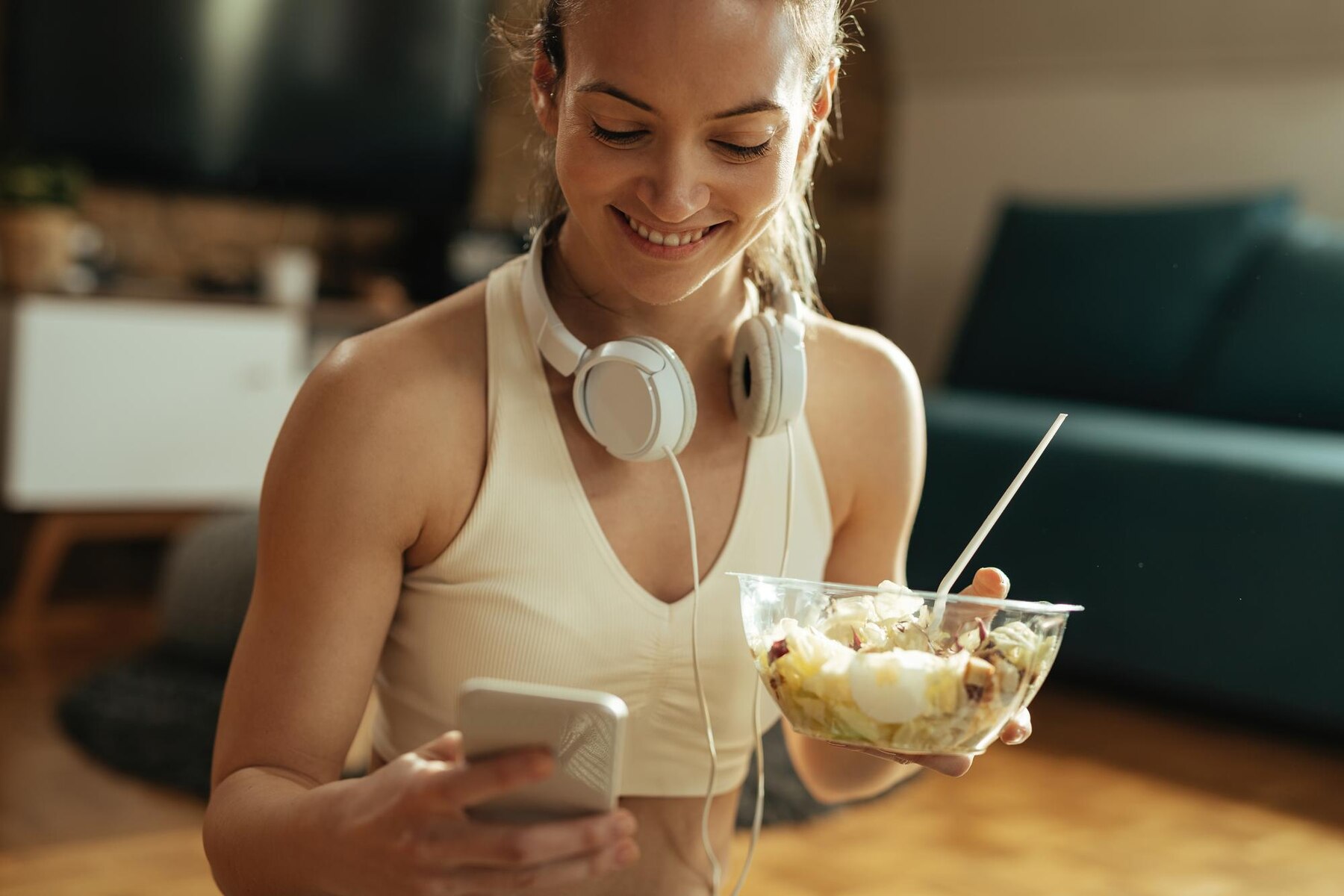 A person wearing a light sports bra sits indoors holding a bowl of salad with a fork in one hand and a smartphone in the other. Over-ear headphones rest around their neck, and the environment appears to be a cozy living room with a sofa and wooden floor in the background. The mood is casual and relaxed, suggesting a break or post-workout meal.