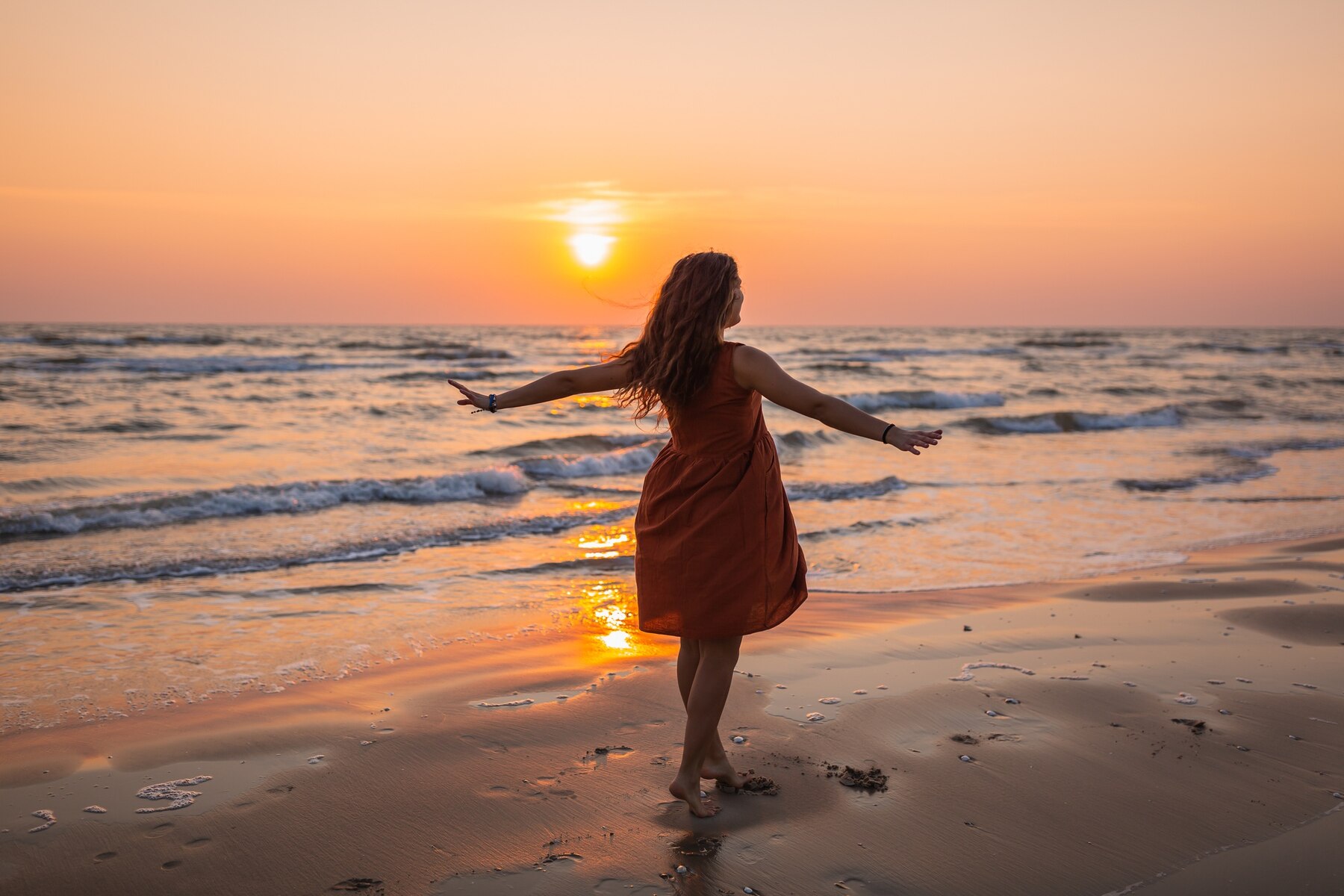 A woman in an orange dress walks barefoot along the shoreline at sunset, arms outstretched as she faces the ocean. Gentle waves roll onto the sandy beach, reflecting the warm golden light of the setting sun. The atmosphere feels peaceful, free, and joyful, evoking a sense of relaxation and connection with nature.