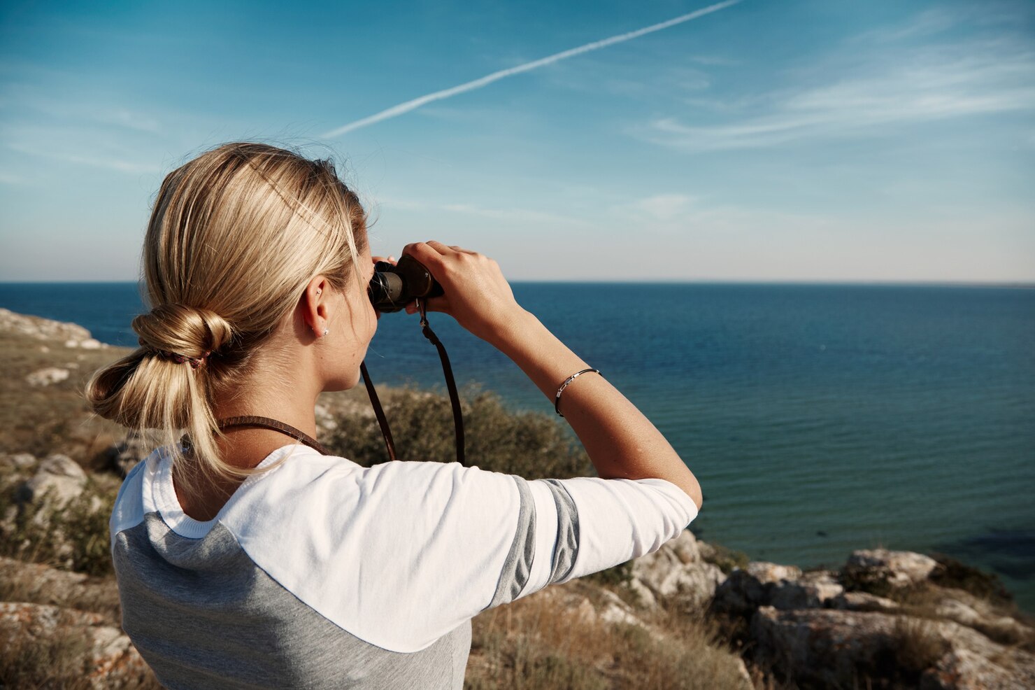 Woman with blonde hair tied back stands on a rocky hillside overlooking the ocean, holding binoculars up to her eyes and gazing toward the horizon. The sky is clear and blue with a single white contrail, and the scene feels calm and contemplative, suggesting curiosity and exploration in a peaceful coastal environment.