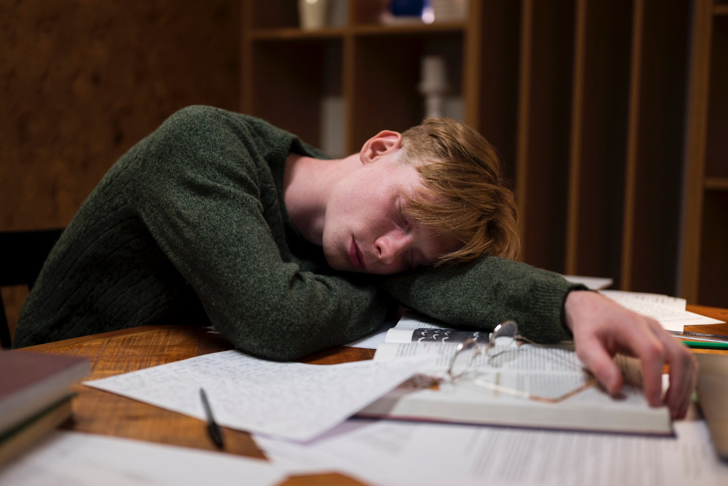 Person wearing a green sweater slumped over a wooden table covered with scattered papers, books, and a pair of eyeglasses, appearing exhausted or overwhelmed. The background features shelves with books and decorative items, and the overall atmosphere suggests fatigue or stress in a study or work environment.