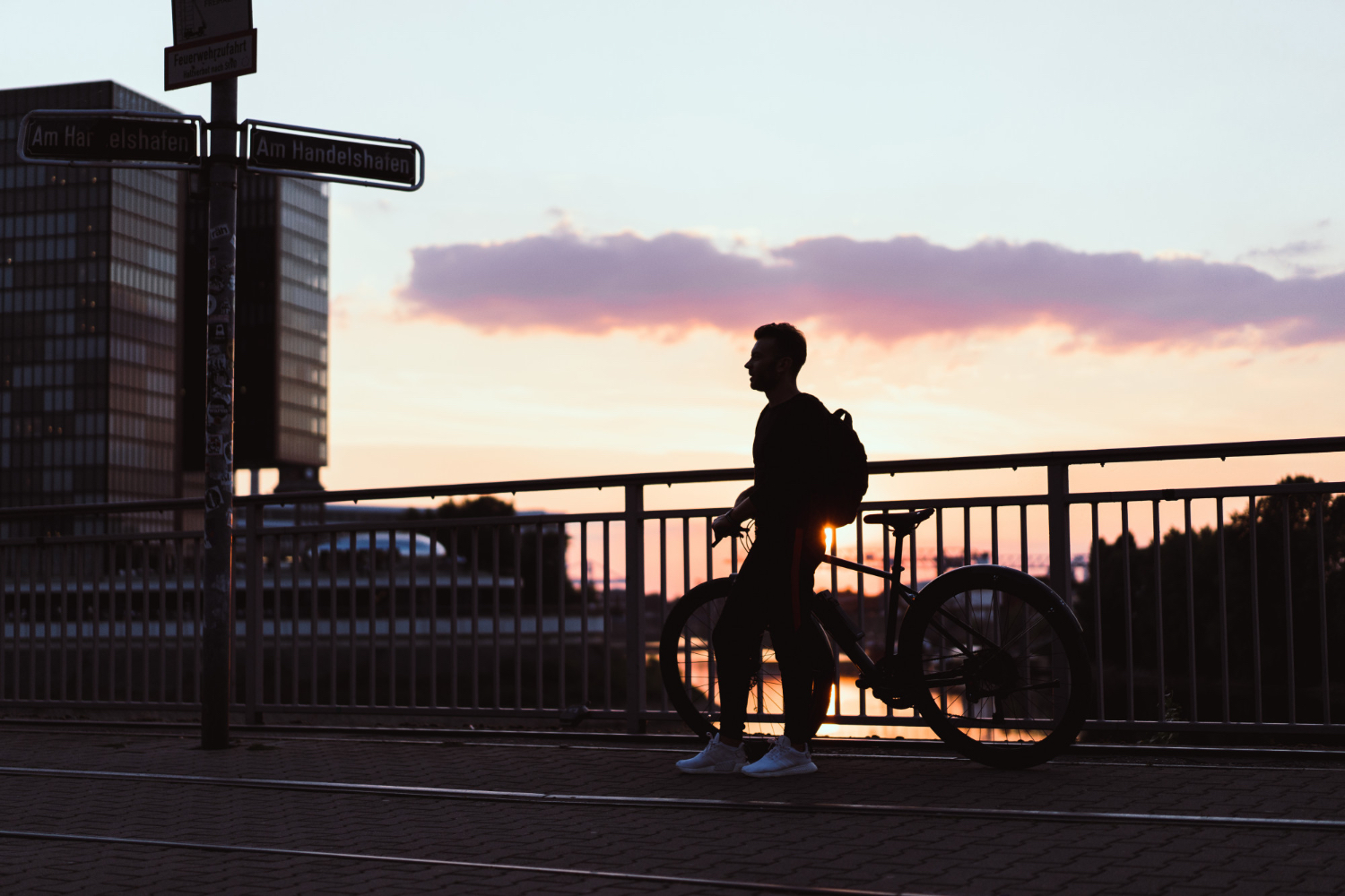 Person with a backpack standing next to a bicycle on a bridge at sunset, silhouetted against a colorful sky with pink and orange clouds. Modern office buildings and street signs are visible in the background, and the overall mood is calm and contemplative.