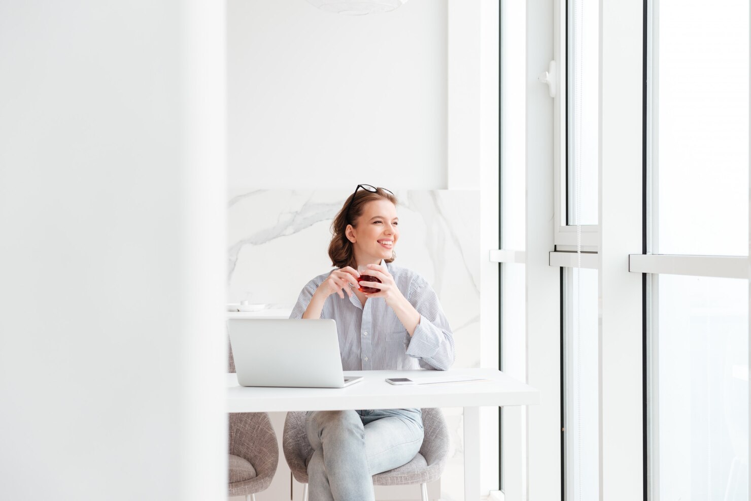Person with shoulder-length hair sitting at a white table in a bright, modern room with large windows, holding a cup with both hands and looking forward. A laptop and smartphone are on the table, suggesting a work or study setting. The atmosphere is calm, minimalistic, and filled with natural light.