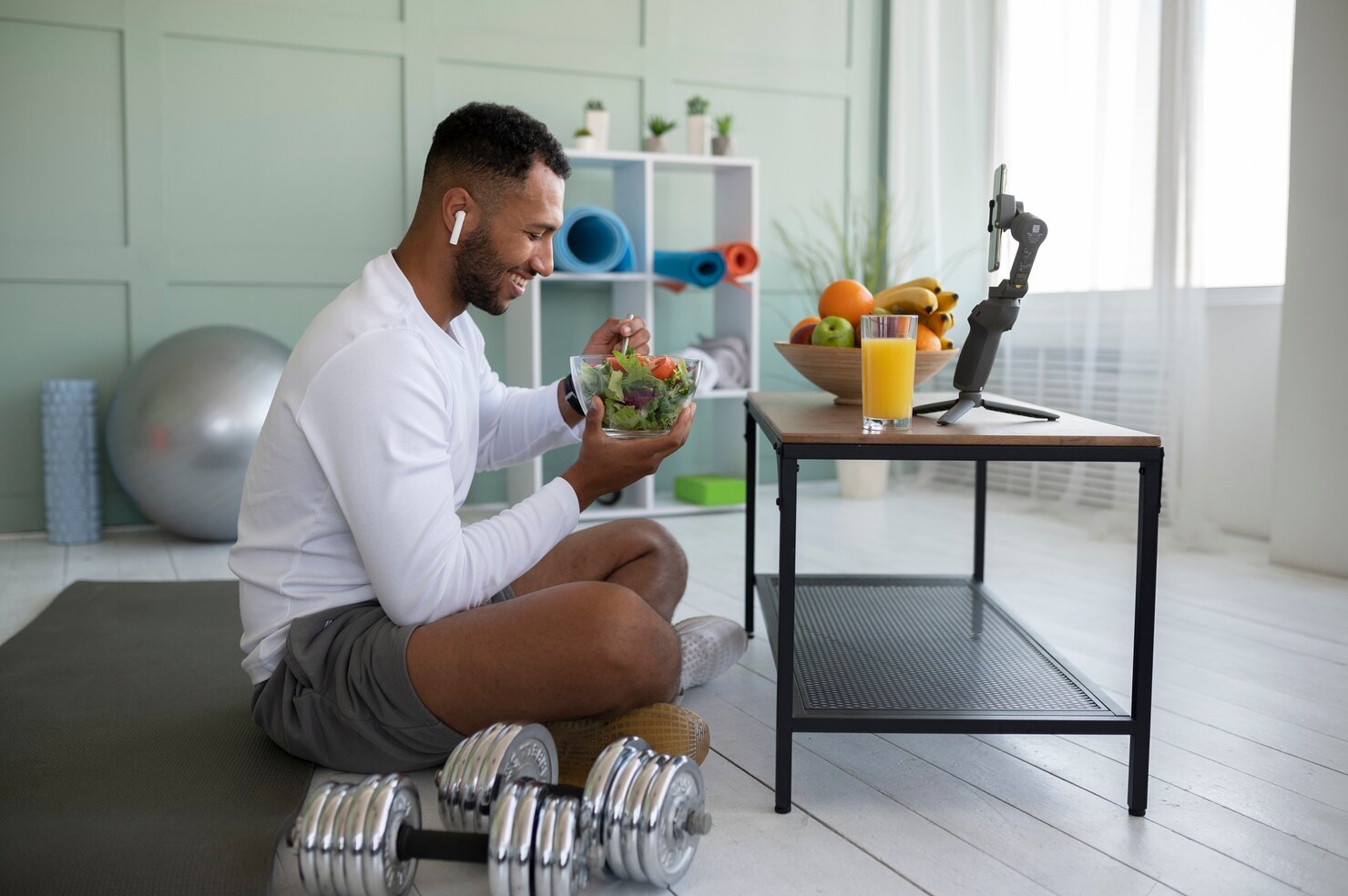 Man in athletic wear sitting cross-legged on a yoga mat in a bright, modern room, holding a bowl of salad and eating with a fork. A glass of orange juice and a smartphone on a tripod are on a table in front of him, along with a bowl of fruit. Dumbbells rest on the floor nearby, and shelves with fitness equipment and plants are in the background. The atmosphere is calm and focused, suggesting a healthy lifestyle and balanced routine.