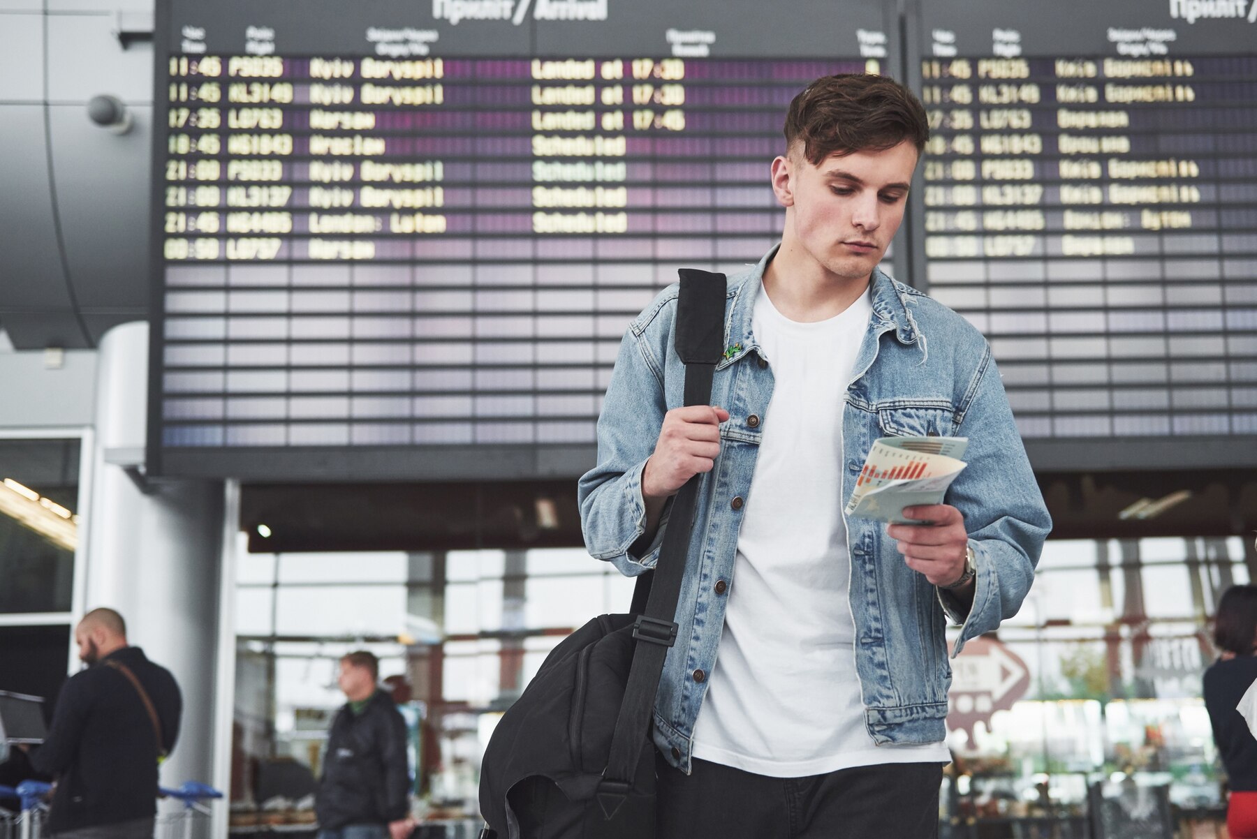 A young man stands in an airport terminal holding travel documents and a black bag, wearing a denim jacket over a white t-shirt. He looks at his boarding pass or ticket while standing in front of a large digital arrivals board displaying flight information in both English and Russian. The environment is busy with other travelers and modern architecture visible in the background. The mood suggests anticipation and focus, typical of travel settings.