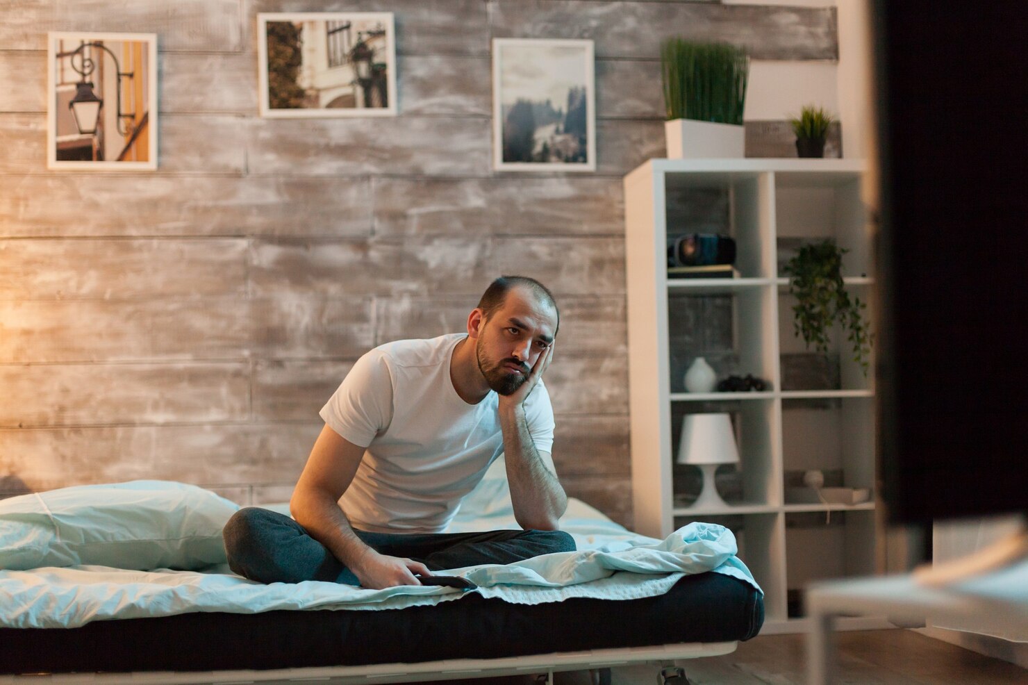 A tired man sits awake on his bed watching television in the middle of the night.