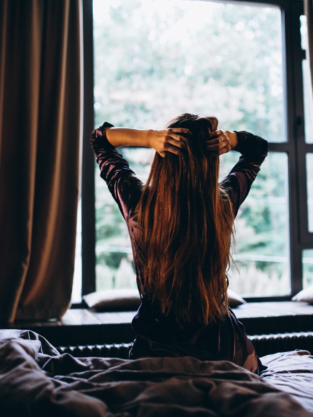 A woman sits on her bed in a dark room framed by heavy opaque curtains and a bright daytime window.