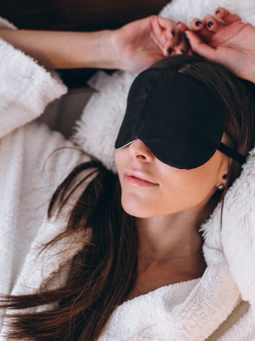 A woman rests in bed wearing a black sleep mask to block light from reaching her eyes.