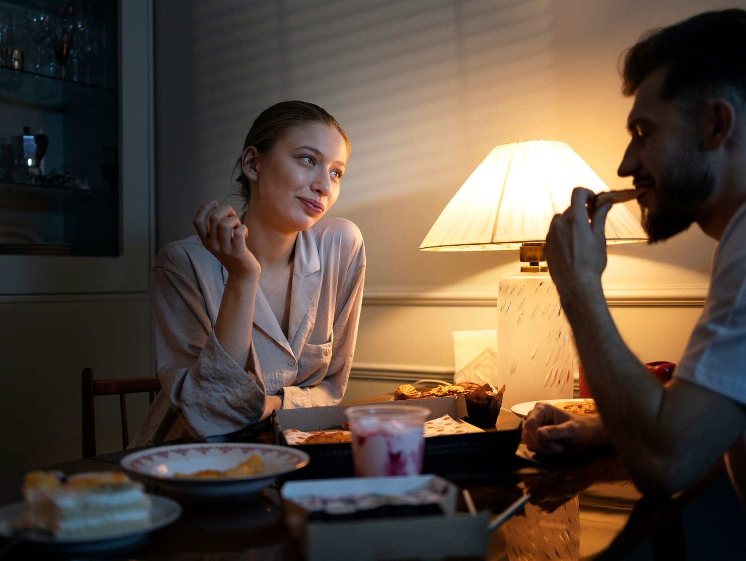 Two people in sleepwear share a late-night meal at a table illuminated by one warm lamp.