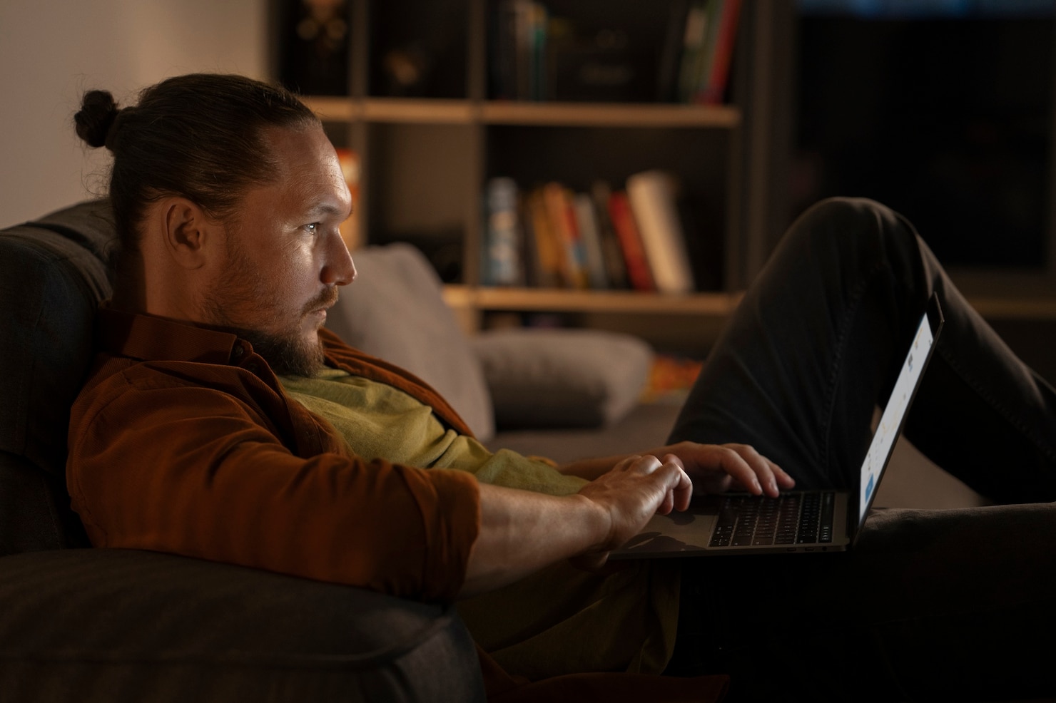 A man remains focused on a laptop in a dark living room late at night.