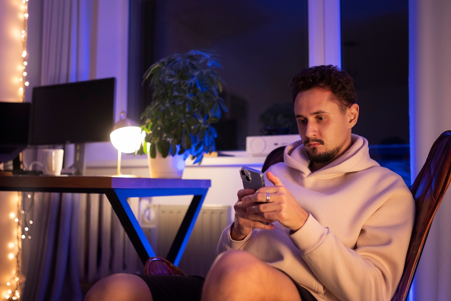 A man looks at his phone beside a dark monitor in a room illuminated by blue window light and bright decorative lights.