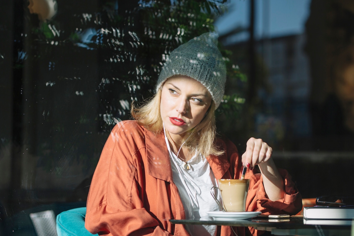 A woman stirs a cup of coffee at a cafe table in strong daytime window light.