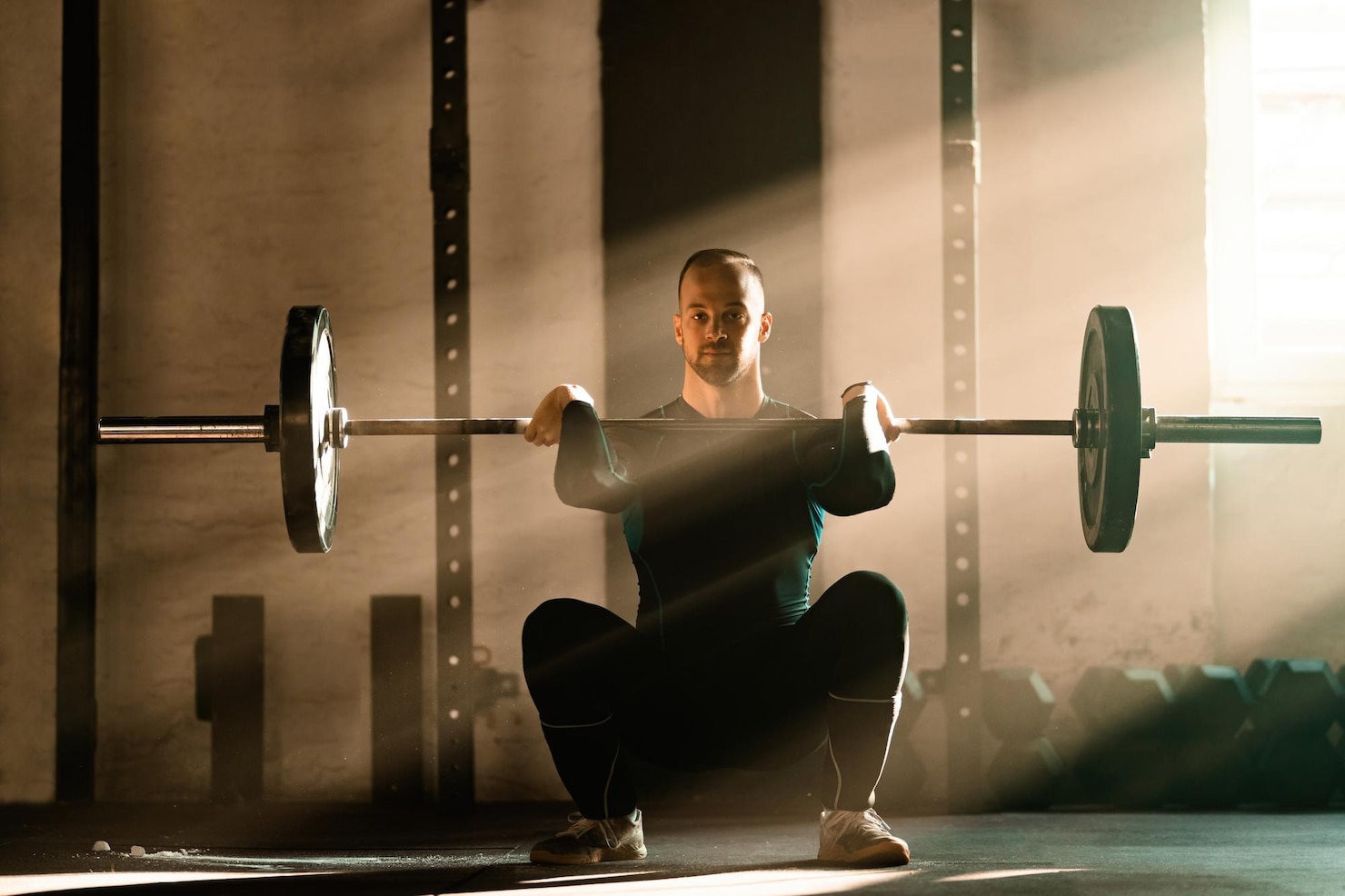 An athlete lifts a barbell in a gym crossed by strong daytime sunbeams.