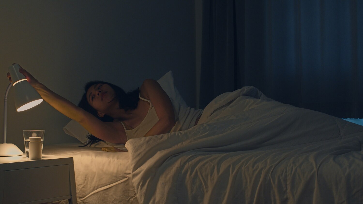 A woman lies awake in a dark bedroom, reaching toward the warm bedside lamp beside her.
