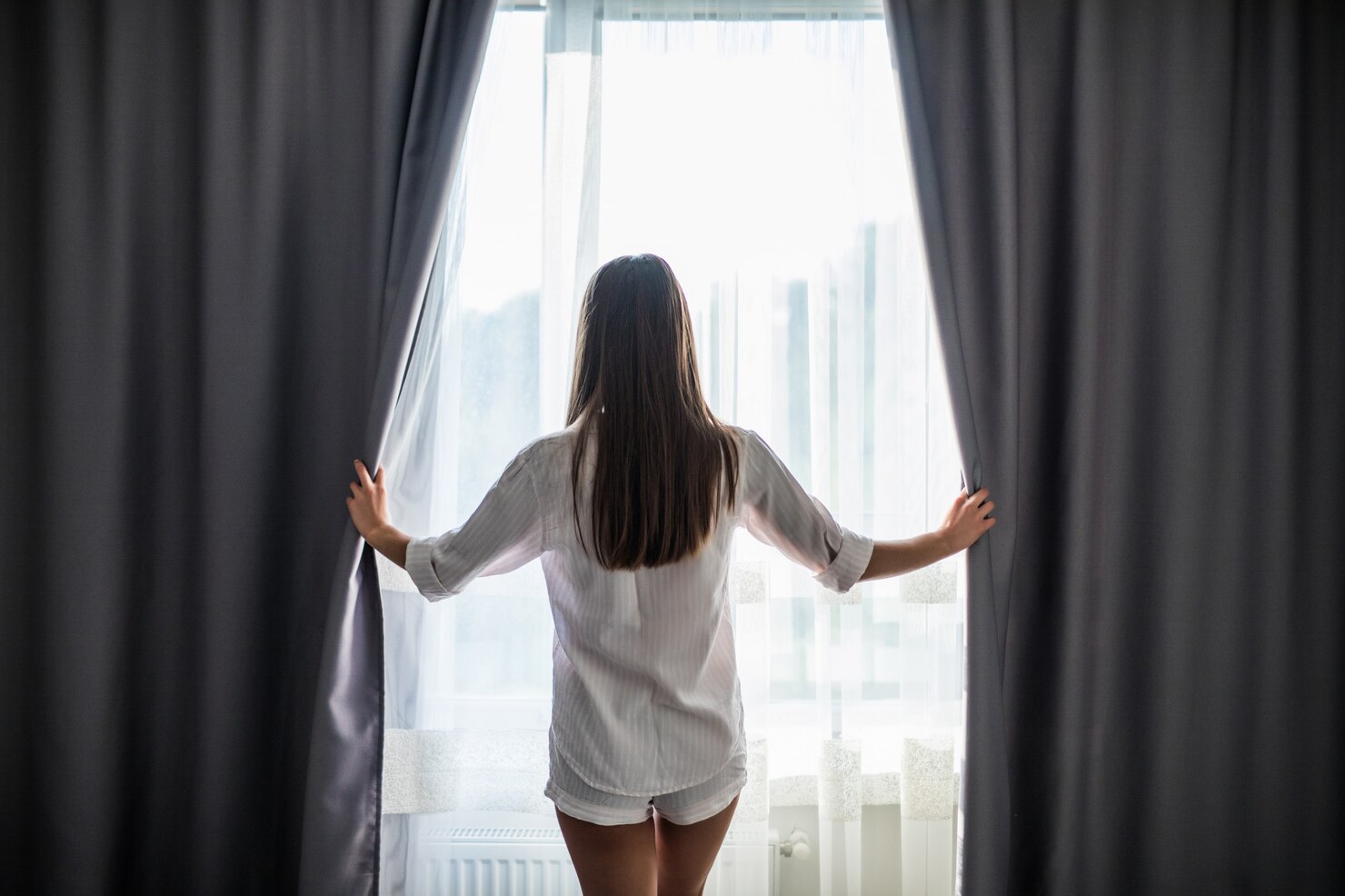 A woman opens dark bedroom curtains to let bright morning light into the room.