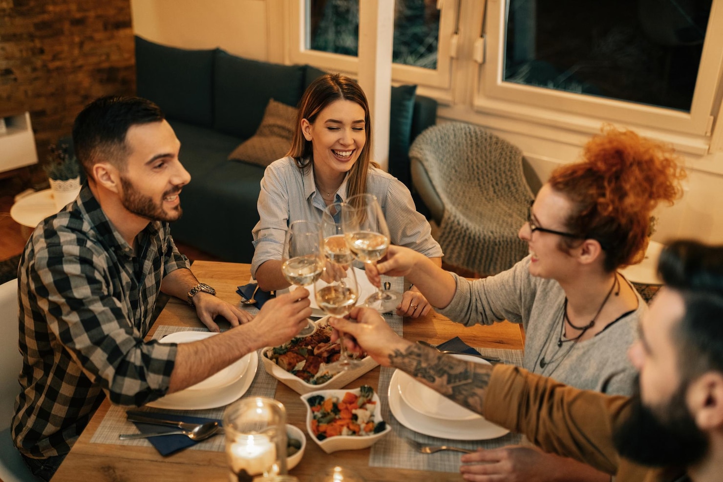 Friends raise glasses around a dinner table during a shared meal in a warmly lit home.