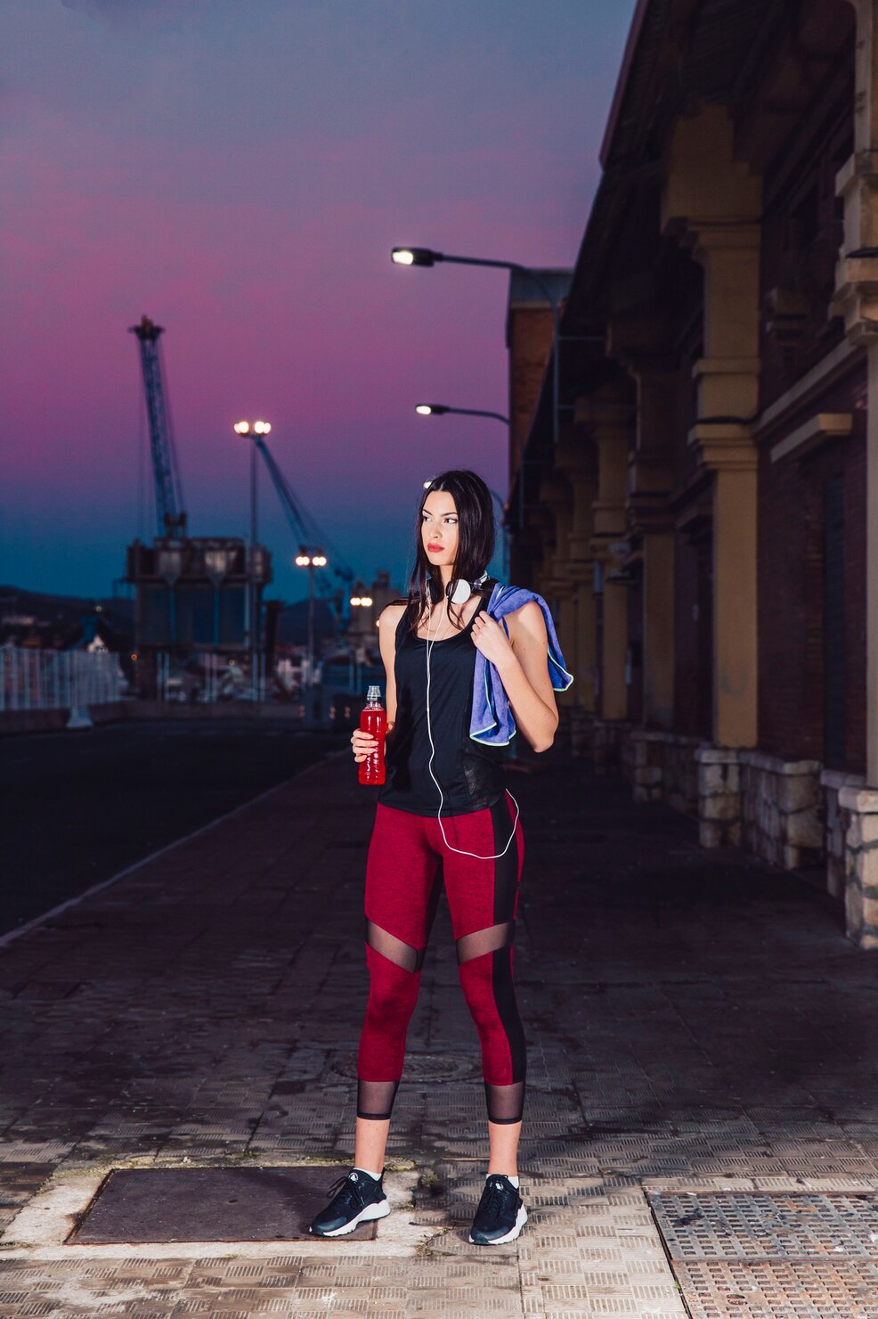 An athlete stands with a towel and drink beneath a violet evening sky in an industrial district.