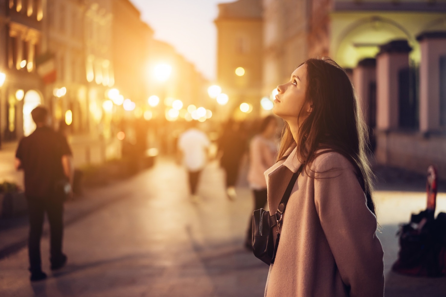 A woman stands on a city street in strong low-angle evening light as the street lamps begin to glow.