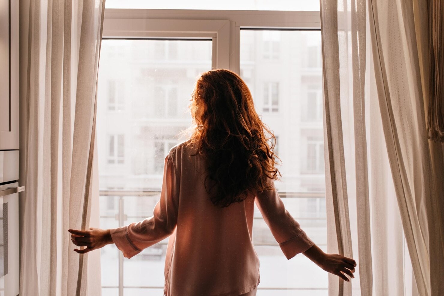A woman opens the curtains as warm morning light fills the room.
