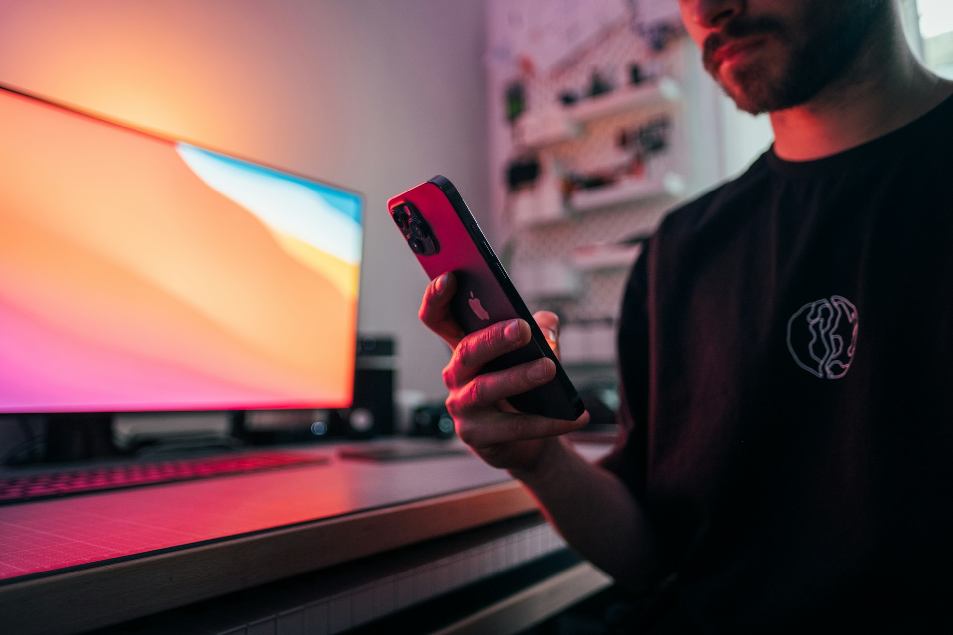 A man with a beard sits at a desk in a softly lit room, holding a smartphone and looking at its screen. A large computer monitor displays a colorful gradient, casting a warm glow over the workspace. The environment feels modern and focused, suggesting concentration or engagement with technology.