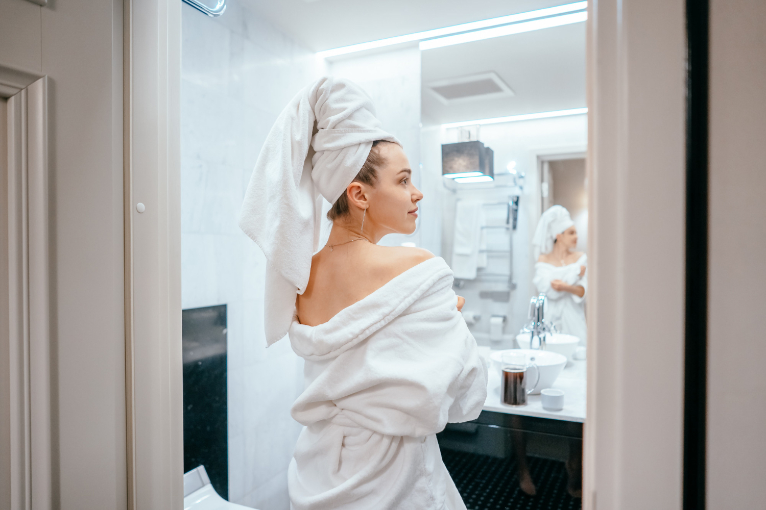 A person wearing a white bathrobe and towel wrapped around their head stands in a modern bathroom, facing a mirror. The bathroom features marble walls, a towel rack, a white sink with a coffee mug, and bright overhead lighting. The atmosphere feels calm and relaxed, suggesting a morning or self-care routine.