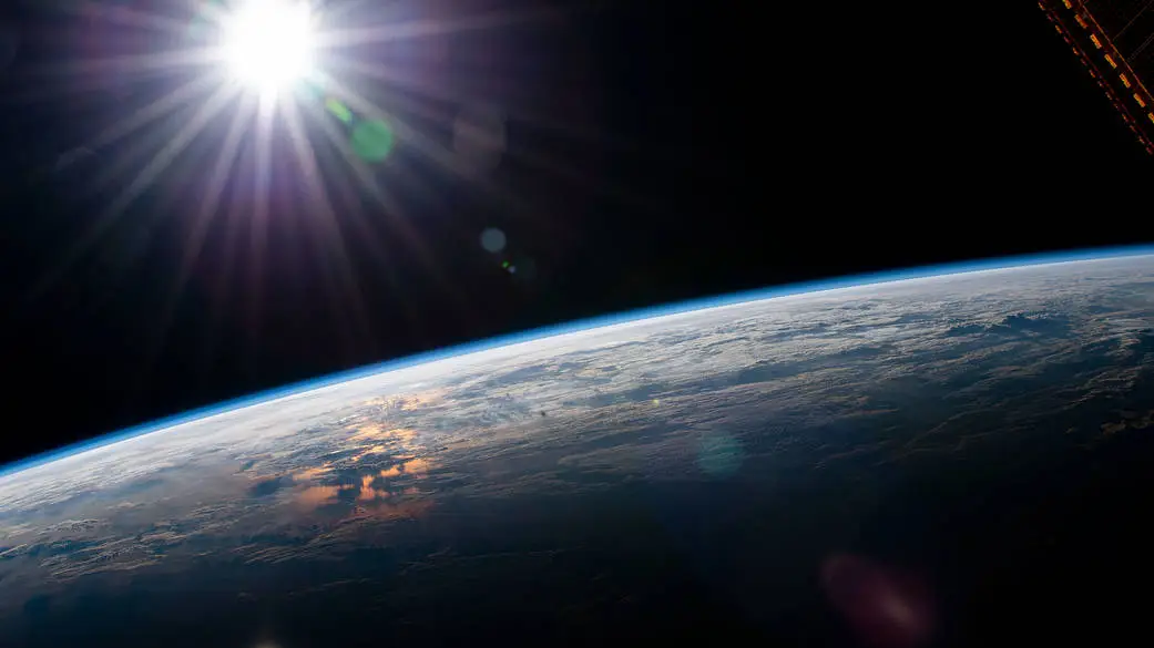 View of Earths curved horizon from space with the sun shining brightly above, casting rays and lens flares across the dark sky. The planet below is partially illuminated, showing clouds and ocean reflecting sunlight. The scene feels awe-inspiring and serene, emphasizing the vastness and beauty of space.