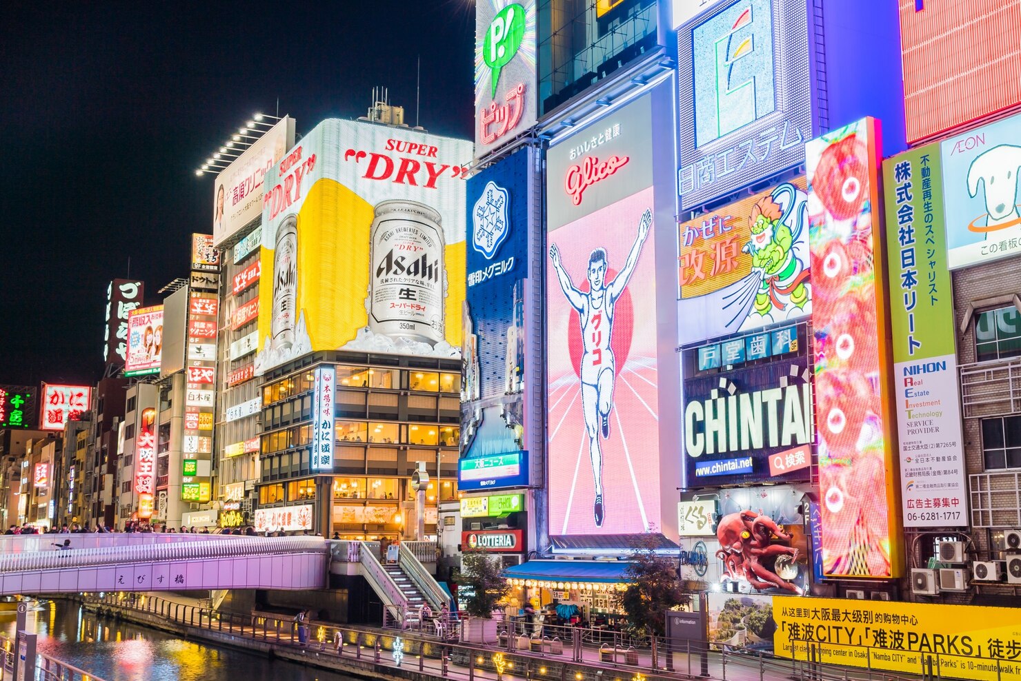 Colorful neon billboards and advertisements illuminate a busy urban street at night in Dotonbori, Osaka. The scene features the iconic Glico running man sign, a large Asahi Super Dry beer ad, and various other vibrant displays with Japanese text. Reflections from the lights shimmer on the canal below, and people walk along the riverside promenade, creating an energetic and lively city atmosphere.