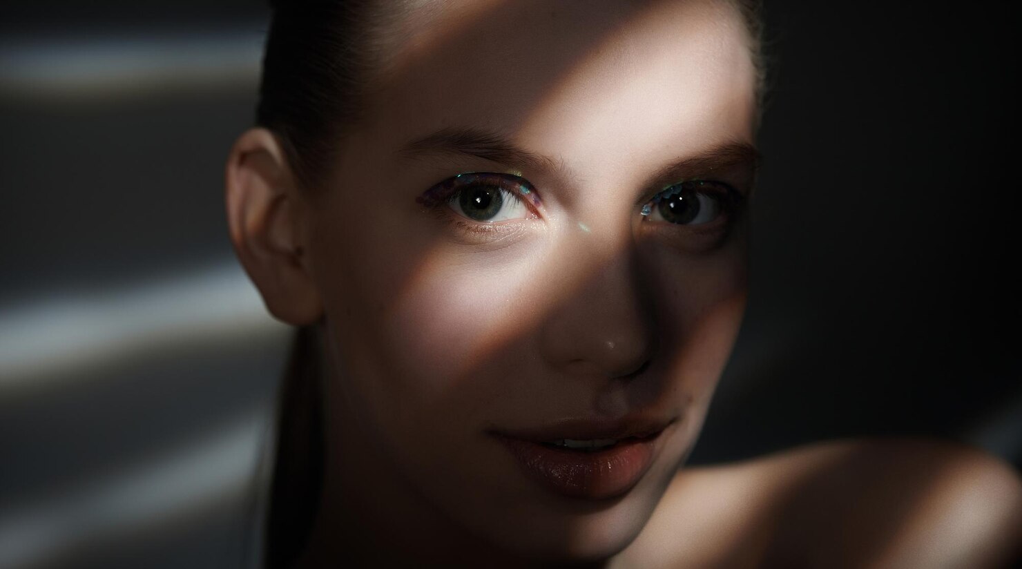 Person with dark hair pulled back, visible ear, and bare shoulder in the foreground, set against a softly lit background with blurred light and shadow patterns. The environment feels calm and intimate, with a focus on quiet reflection or solitude.
