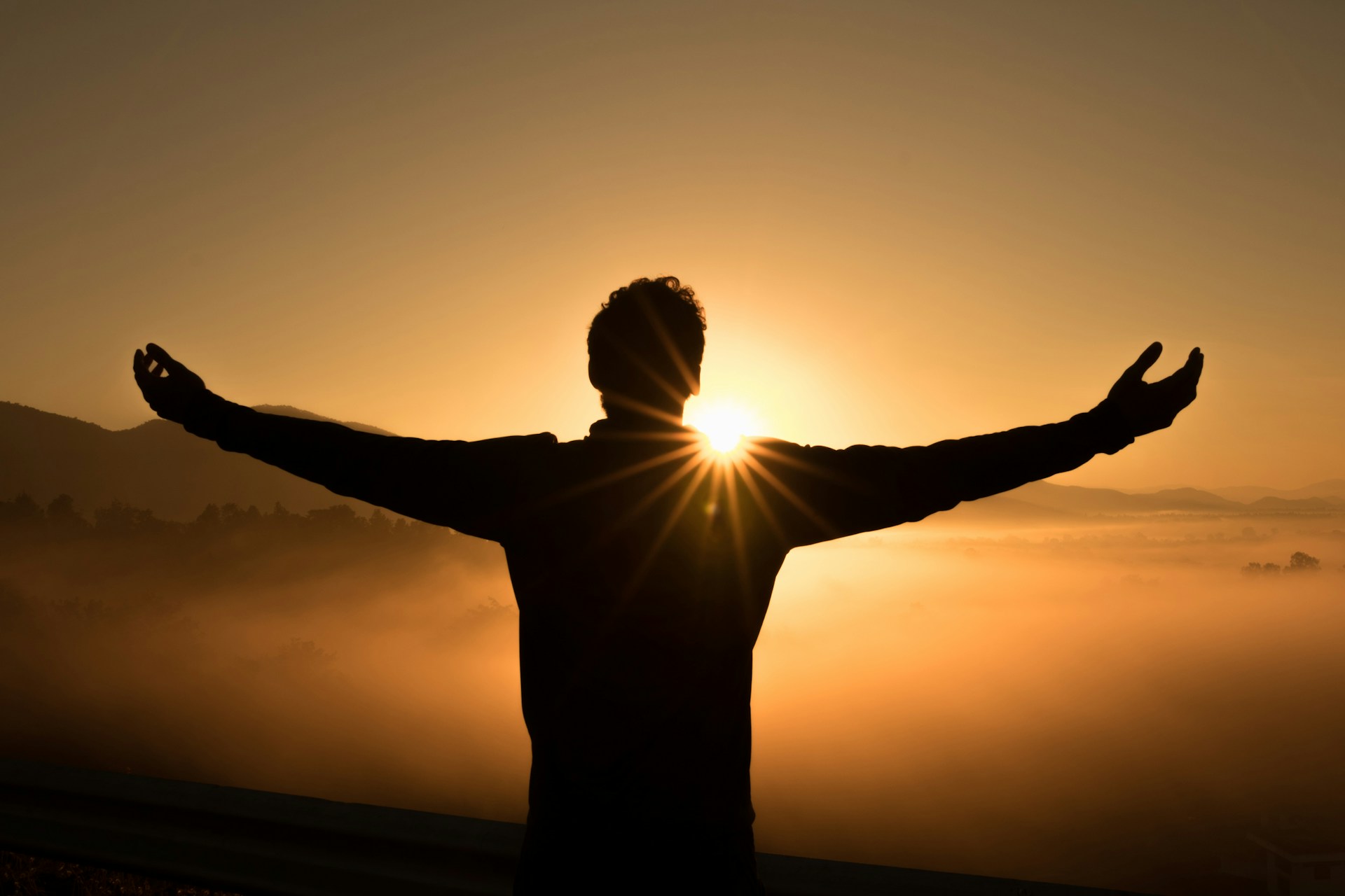 Silhouette of a person standing outdoors at sunrise with arms outstretched, facing the rising sun. The sun is partially obscured by the person's body, creating a radiant glow and sunbeams. The background features misty hills and a golden sky, evoking a sense of freedom, hope, and tranquility.