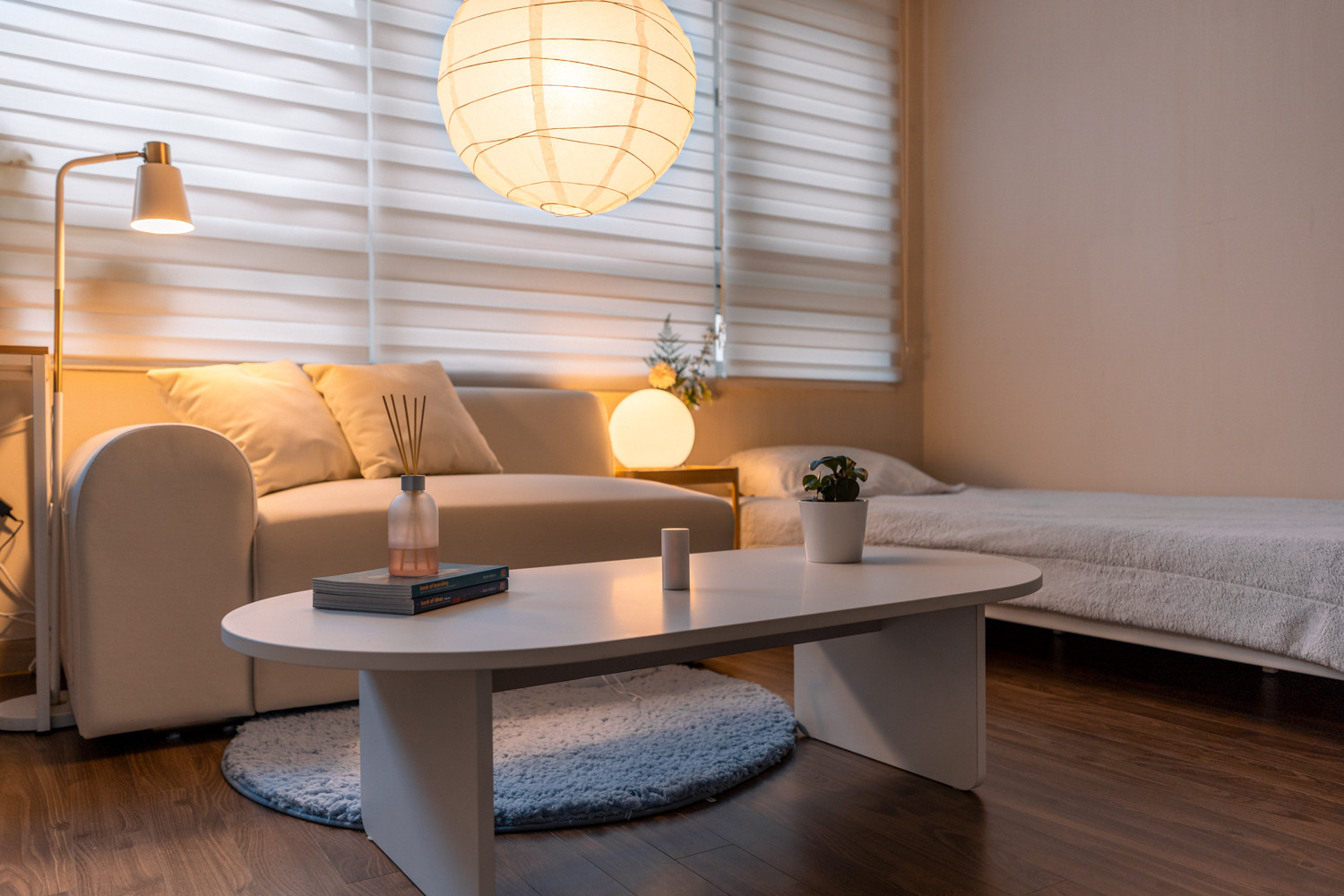 A minimalist living space featuring a white sofa with plush pillows, a low white coffee table holding books, a reed diffuser, and a small potted plant. A neatly made bed with light bedding sits nearby, and the room is softly illuminated by a large paper lantern and natural light filtering through white blinds. The environment feels calm, cozy, and organized, evoking a sense of peaceful relaxation.