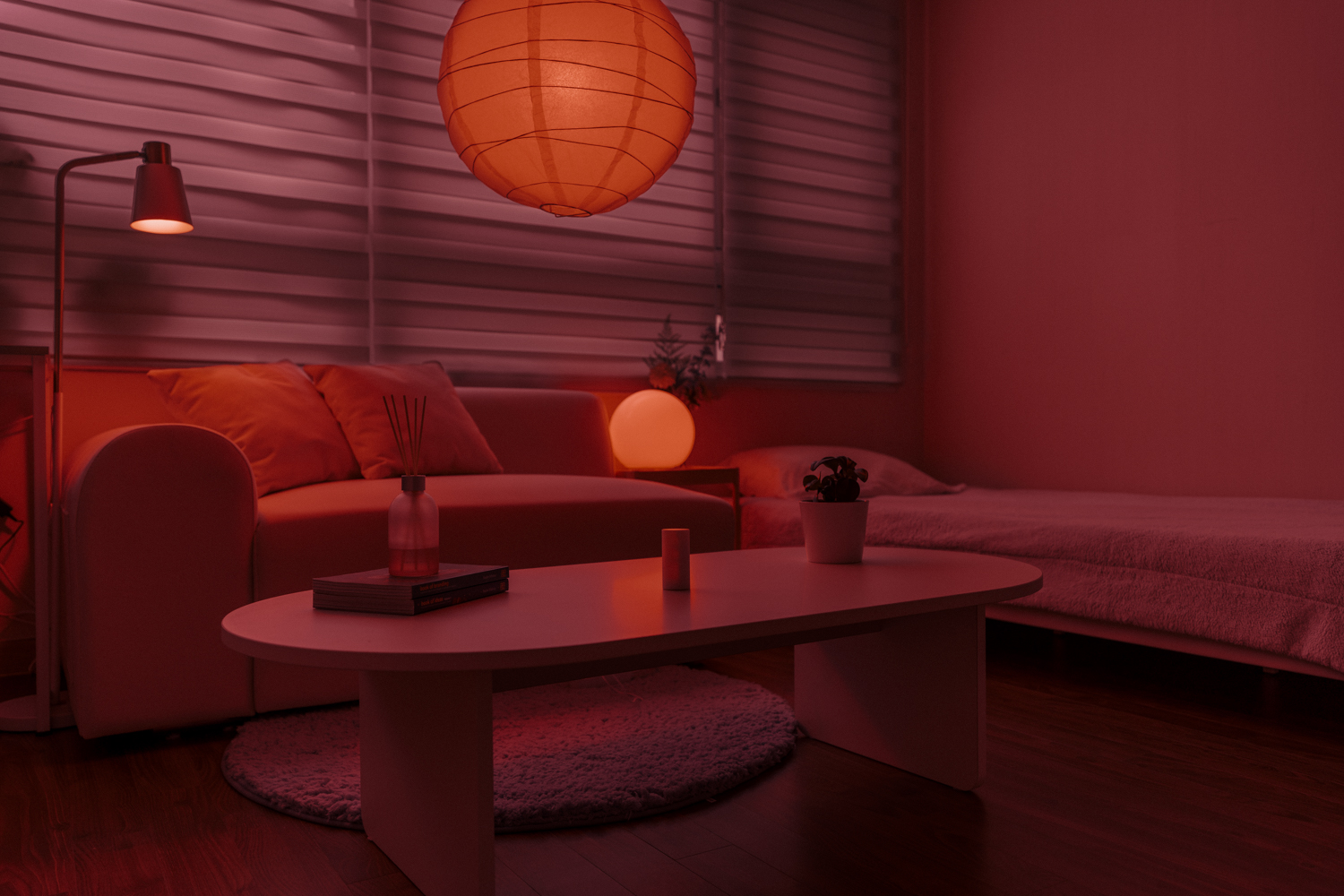A minimalist living room with a white sofa and plush pillows sits beside a low white coffee table holding books, a reed diffuser, and a small potted plant. A neatly made bed with light bedding is nearby. The space is softly illuminated by a large paper lantern and warm red ambient lighting, with natural light filtering through white blinds. The environment feels calm, cozy, and organized, evoking peaceful relaxation.