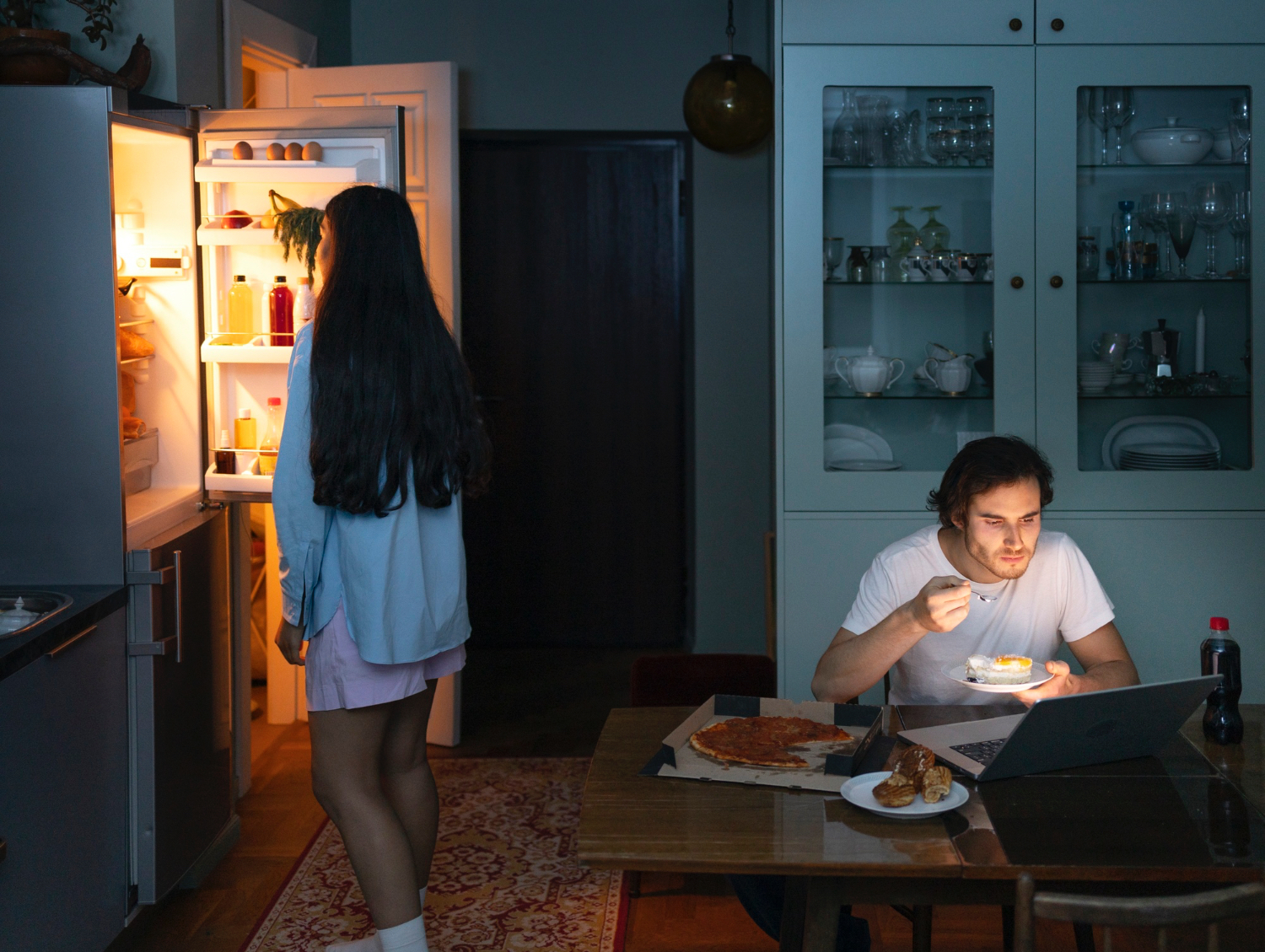Woman with long dark hair stands in front of an open refrigerator, illuminated by its light, while a man sits at a dining table eating from a plate and looking at a laptop. The table holds a pizza, pastries, and a bottle of soda. The kitchen and dining area are dimly lit, with cabinets displaying glassware and dishes in the background. The scene feels quiet and relaxed, suggesting a late-night meal or snack at home.