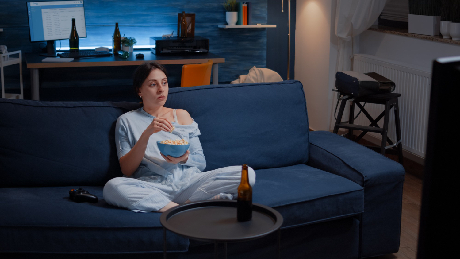 Woman in light blue pajamas sits cross-legged on a dark blue sofa, holding a bowl of popcorn and watching television. A beer bottle rests on a round coffee table in front of her, and a game controller is on the sofa nearby. The room is dimly lit with a cozy, relaxed atmosphere, featuring a desk with a computer and bottles in the background, and a window with curtains to the side.