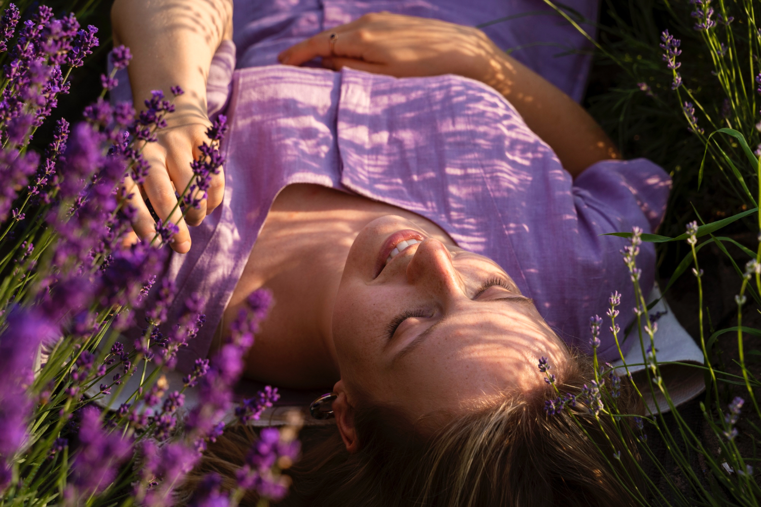 A woman wearing a lavender dress lies on her back in a field of blooming lavender flowers, eyes closed and smiling softly. Sunlight filters through the flowers, casting dappled shadows across her face and body. The scene feels peaceful, relaxed, and immersed in nature, evoking a sense of calm and contentment.