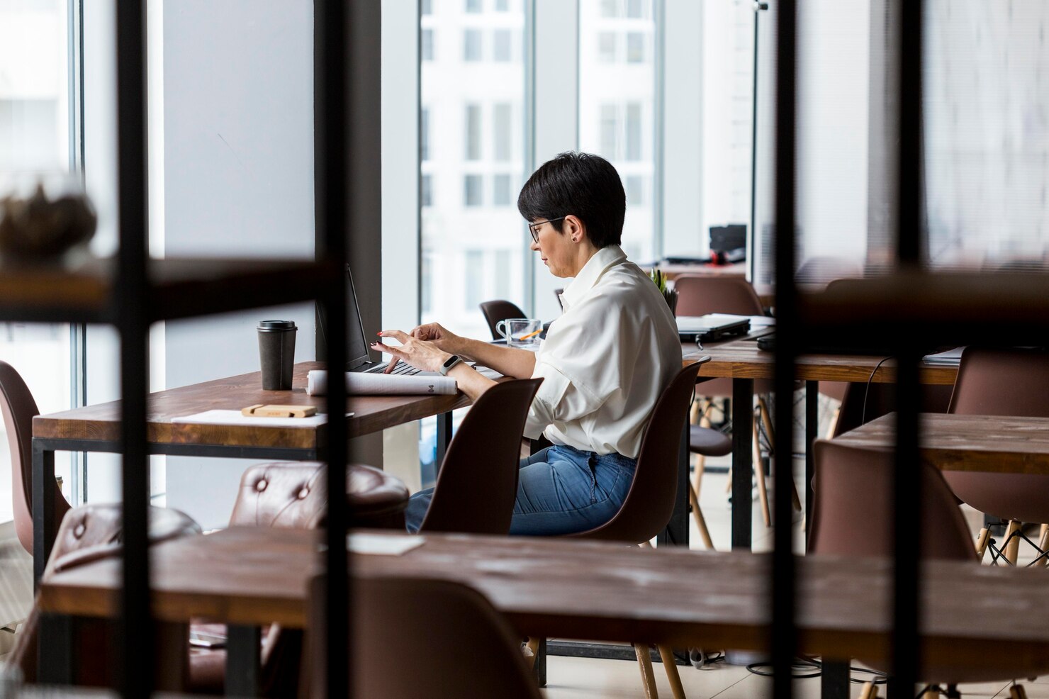 Person with short dark hair sitting at a wooden table in a modern, well-lit cafe, working on a laptop. The person is dressed in a white shirt and blue jeans, focused on the screen with one hand on the keyboard and the other holding a pen. A disposable coffee cup, a glass of water, and a notebook are on the table. The background features large windows with city buildings visible outside, and several empty tables and chairs, creating a calm and productive atmosphere.